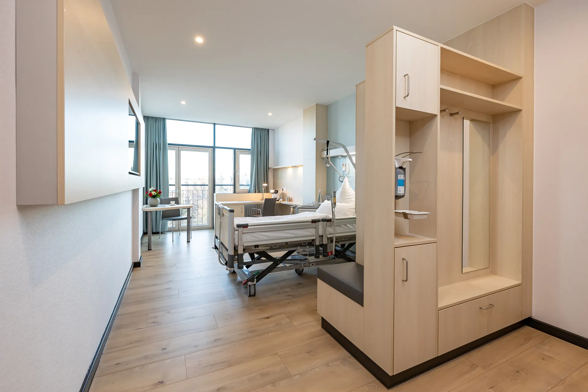 Modern eingerichtetes Patientenzimmer mit Pflegebett, Fensterfront, Schreibtisch und Garderobenschrank
