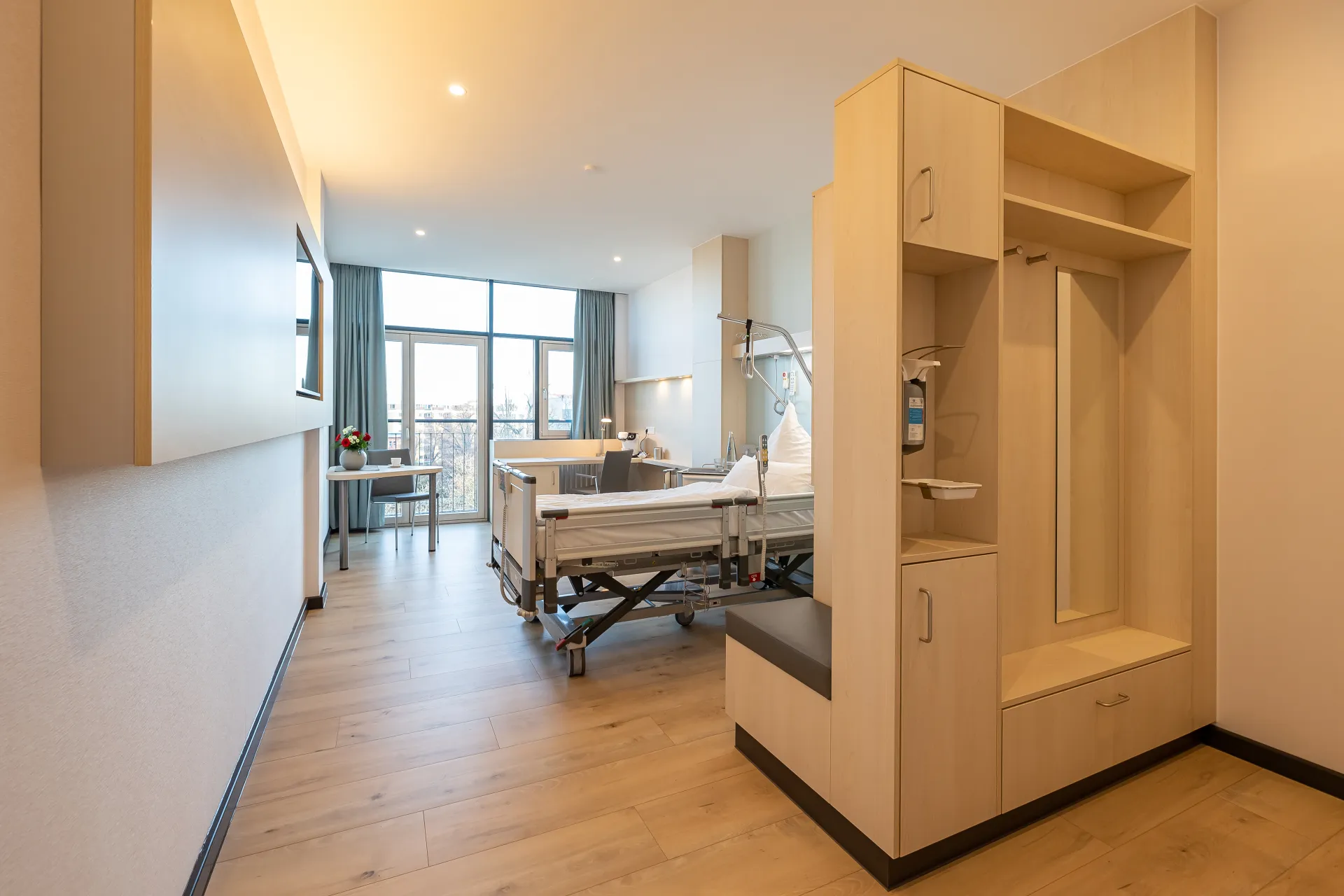 Helles Patientenzimmer mit Pflegebett, Sitzbereich, großem Fenster und Gardinen sowie offenem Schrank mit Sitzbank und Ablagen