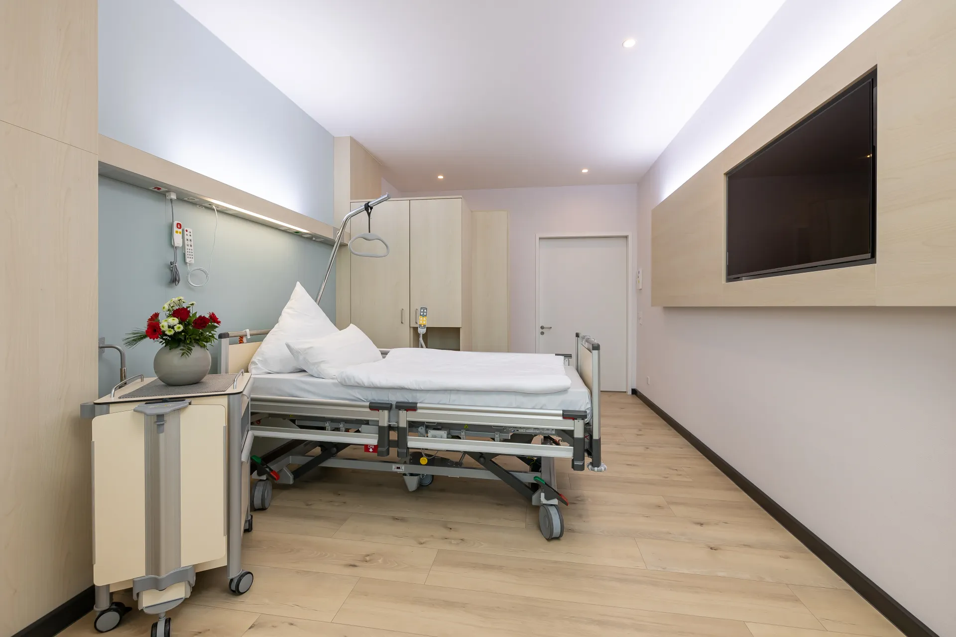 Krankenhauszimmer mit leerem Bett, Nachttisch mit Vase und Blumen, großem Wandfernseher und Holzboden