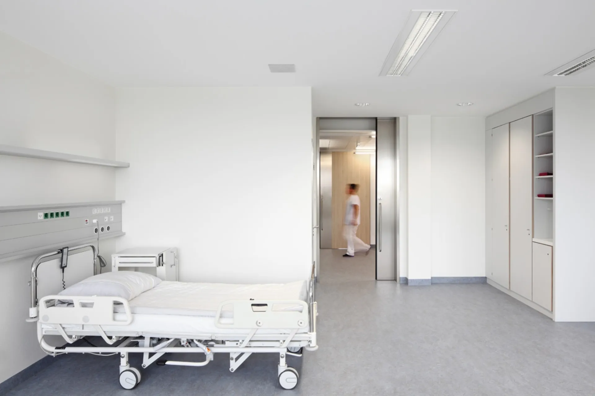 Leerer Krankenhausraum mit einem Bett links und einer Person in weißer Kleidung, die durch eine Tür im Hintergrund geht