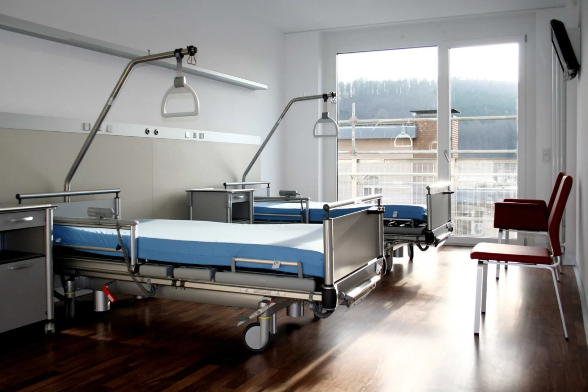 Leerer Krankenhausraum mit zwei Betten, blauen Matratzen, zwei roten Stühlen und großem Fenster mit Blick auf ein Baugerüst und bewaldeten Hügel