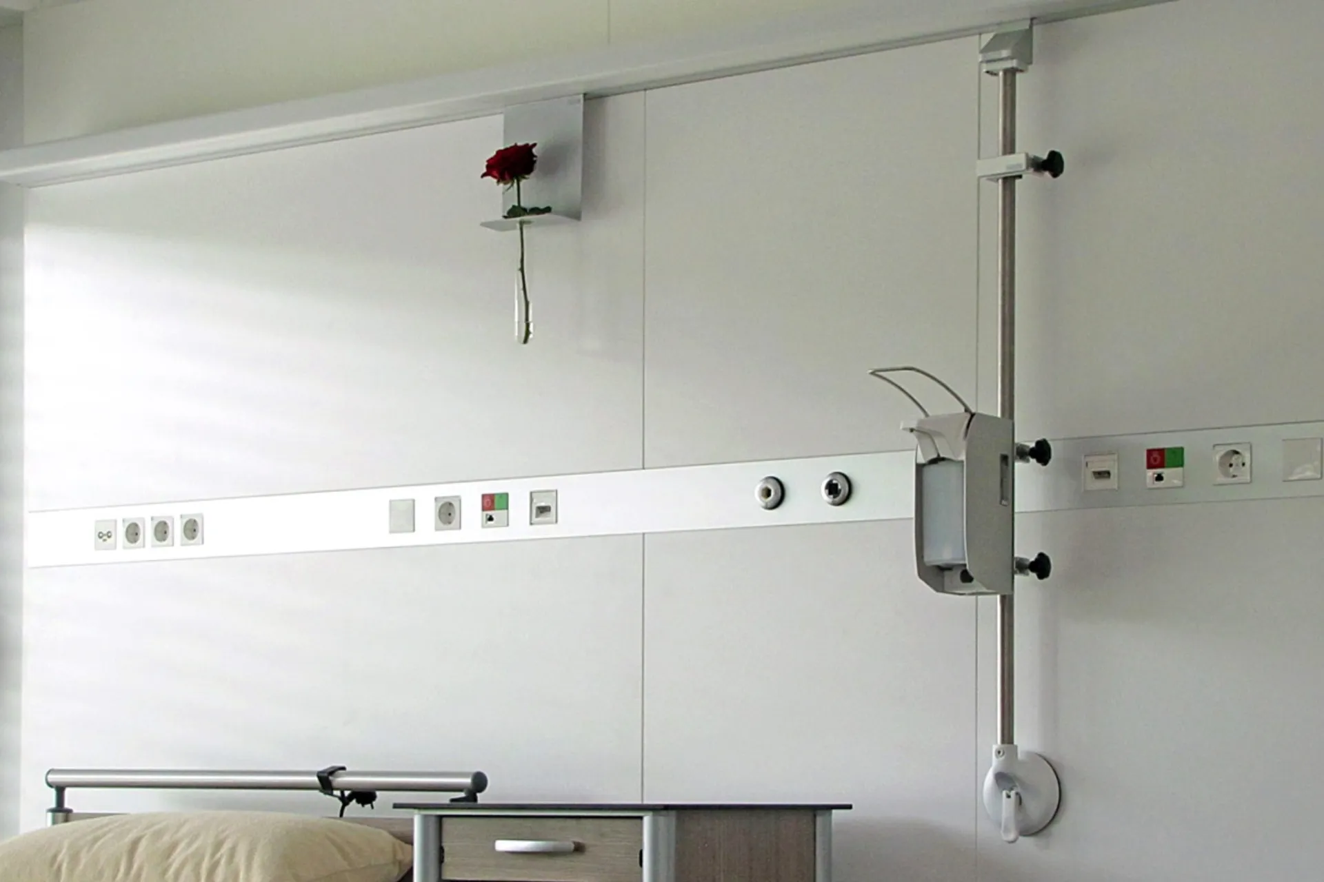 In der horizontalen Geräteschiene an der Rückwand eines Krankenzimmers sind eine Blumenvasenhalterung und eine Stange mit Desinfiziermittel eingehängt.