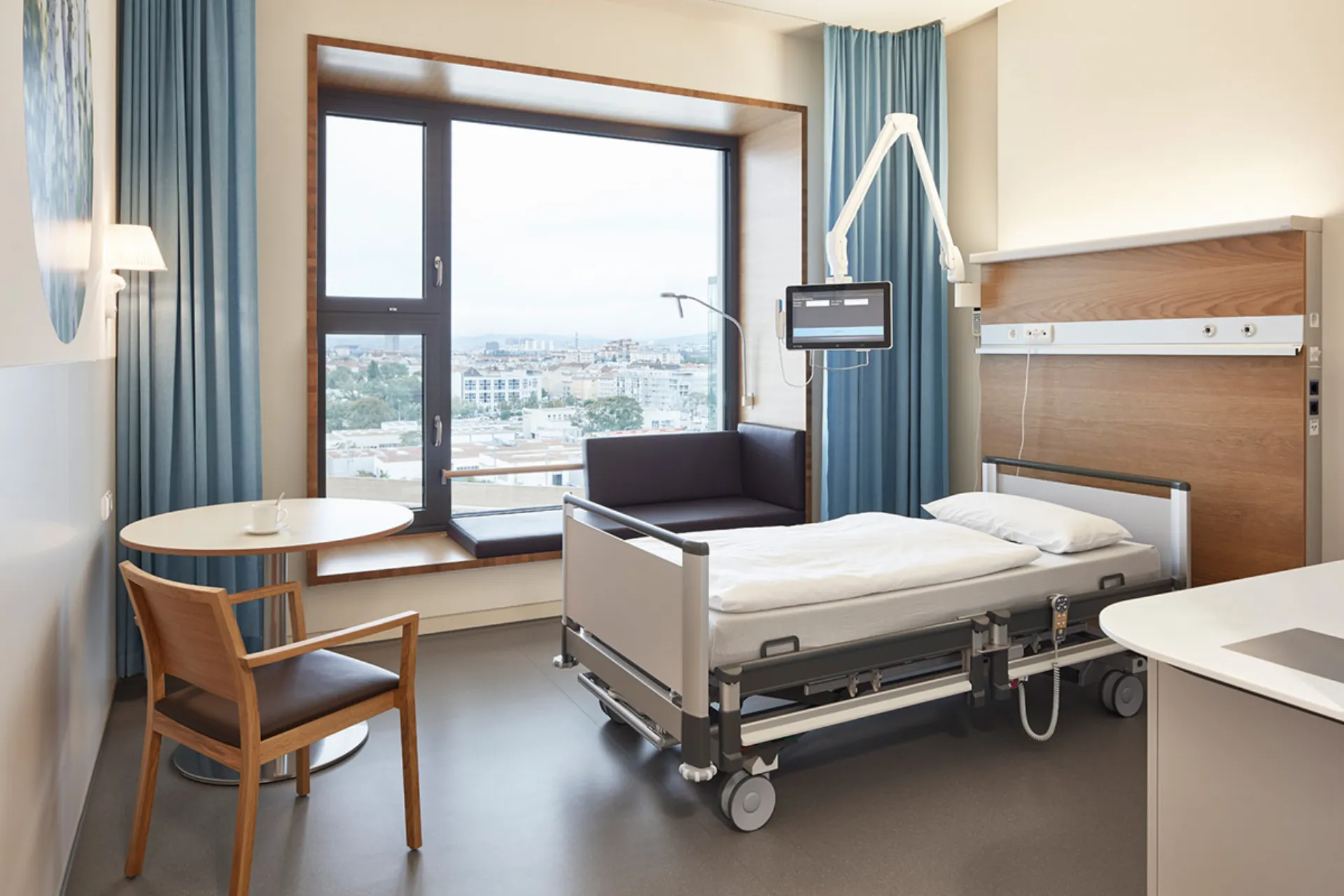 Krankenhauszimmer mit Pflegebett, Fensterbank mit Sitzkissen, Stuhl und kleinem runden Tisch vor großem Fenster mit Blick auf Stadt