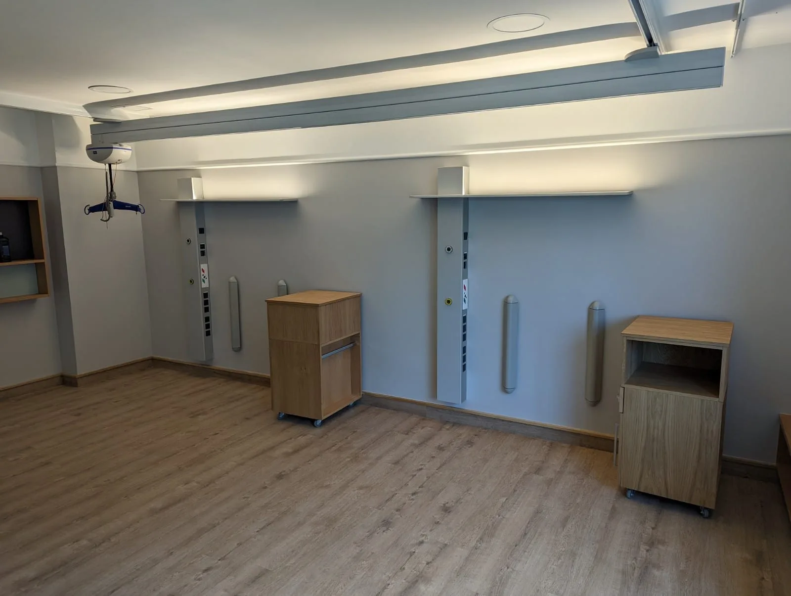 Zweibettbereich mit zwei vertikalen Versorgungssäulen pure an grauer Wand, Holznachttischen, indirekter Deckenbeleuchtung und Patientenlifter
