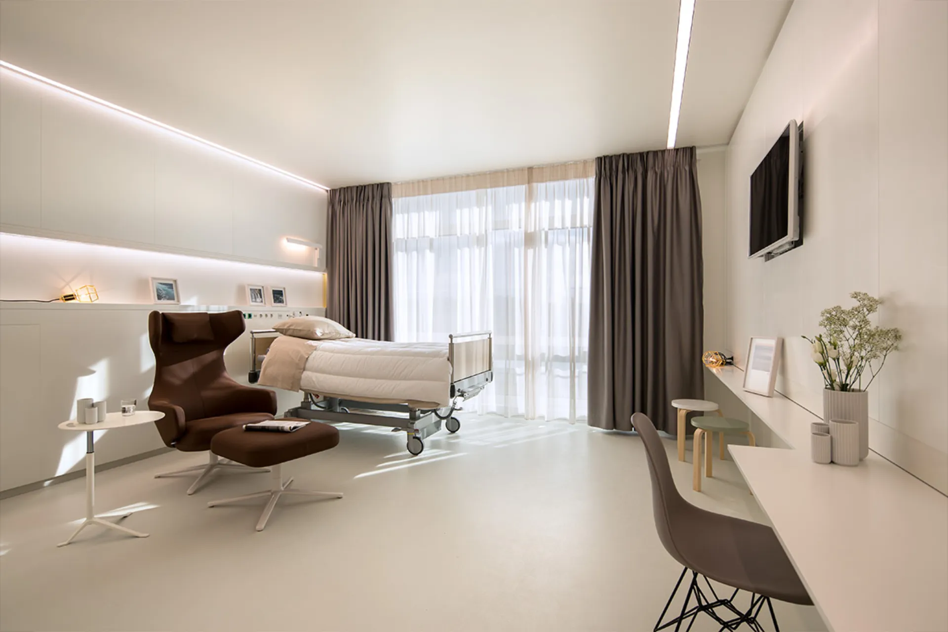 Helles, modernes Patientenzimmer mit Pflegebett, braunem Sessel mit Fußhocker, Schreibtisch und großem Fenster mit Vorhängen