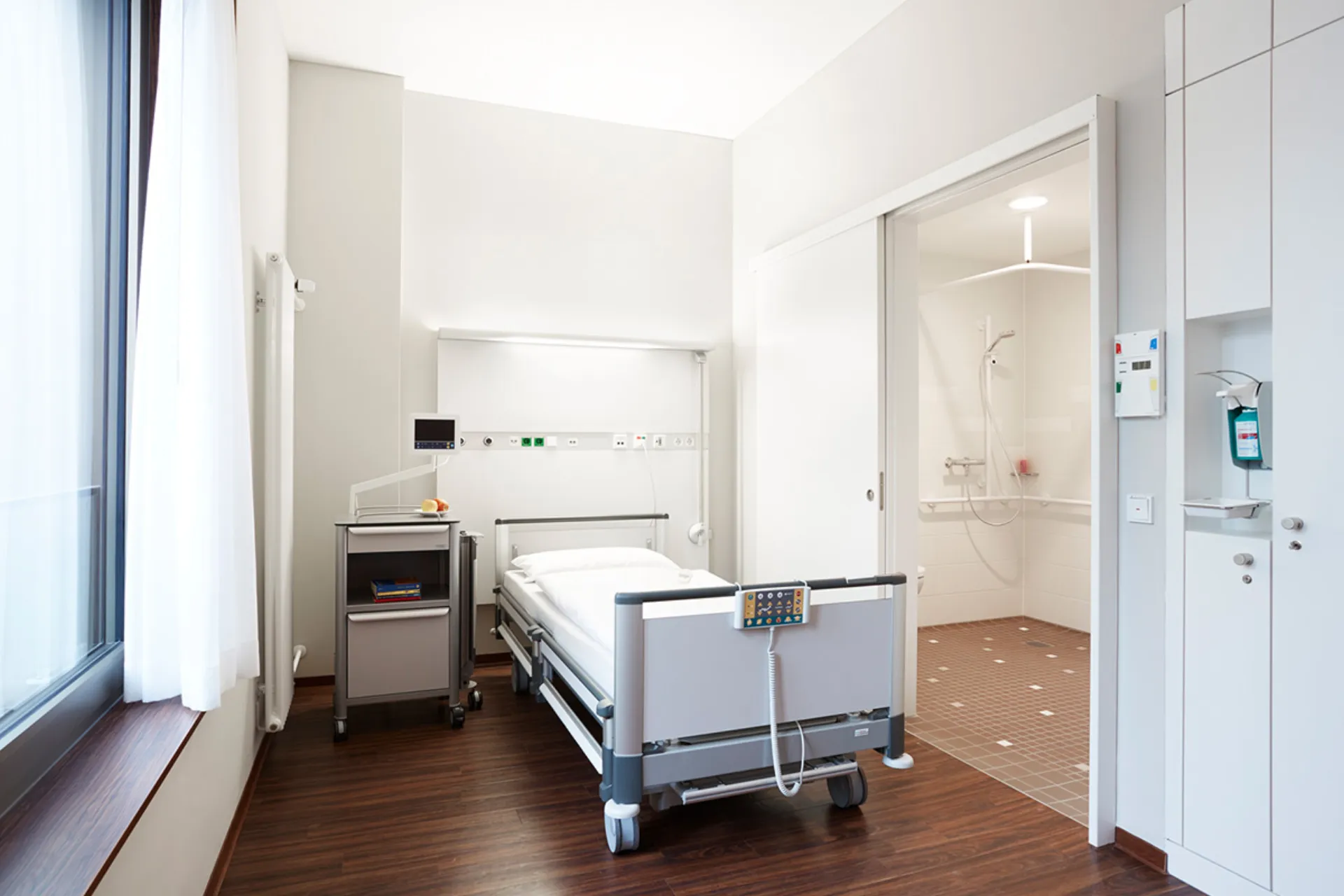 Krankenhauszimmer mit einem Bett, grauer Bettdecke, Nachttisch auf Rollen und weißen Schränken an der Wand