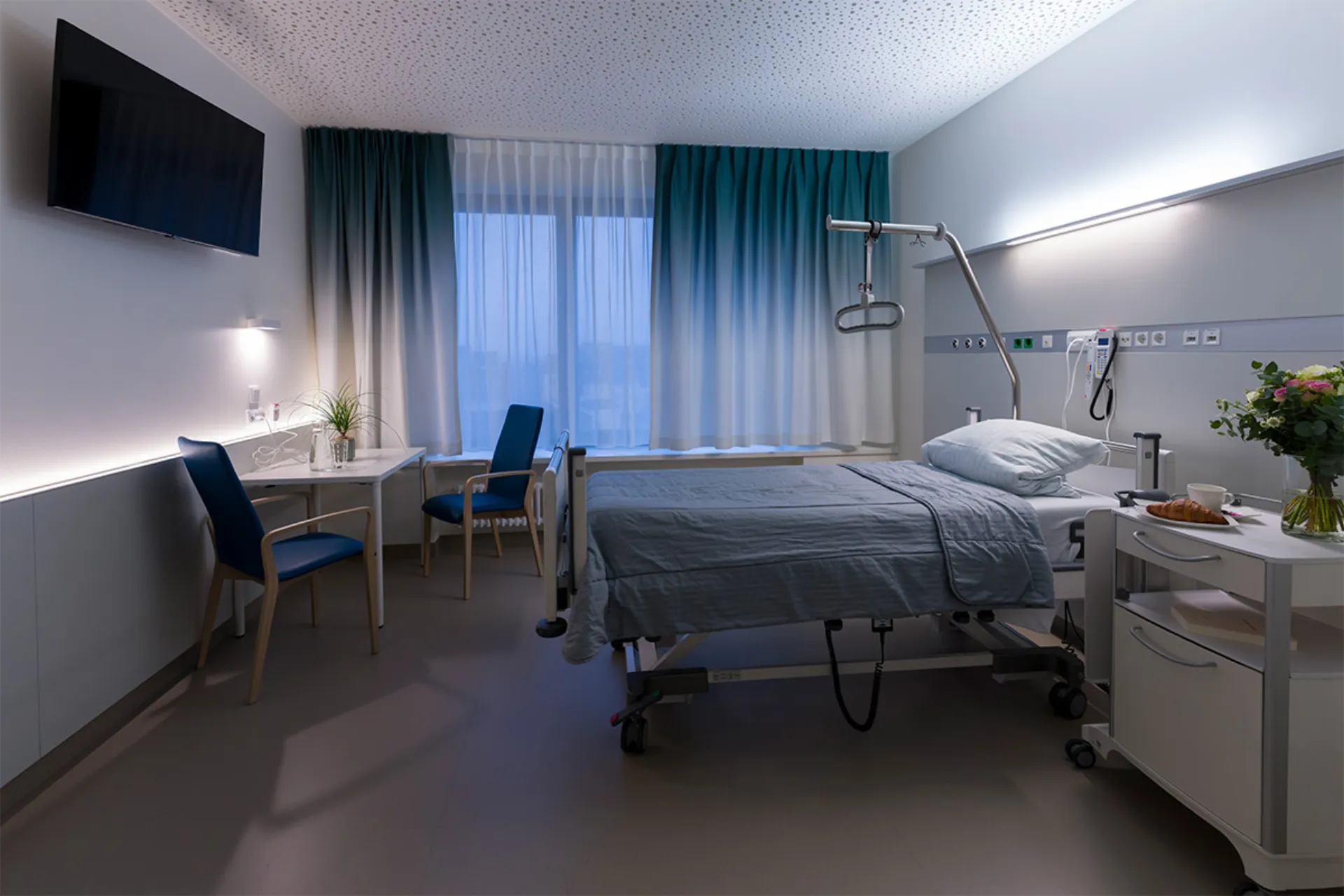 Krankenhauszimmer mit Pflegebett, Nachttisch, zwei Stühlen, Schreibtisch, großem Fenster mit Vorhängen und Wandfernseher
