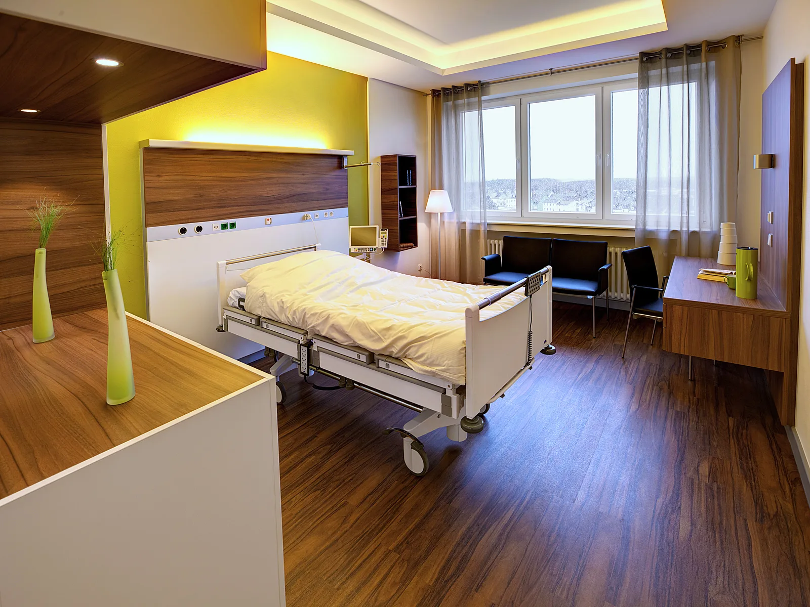 Patientenzimmer mit Pflegebett vor einer Wandverkleidung aus dunklem Holz, gelb hinterleuchteter Versorgungsleiste, Sitzecke mit dunklen Stühlen und großen Fenstern mit Ausblick