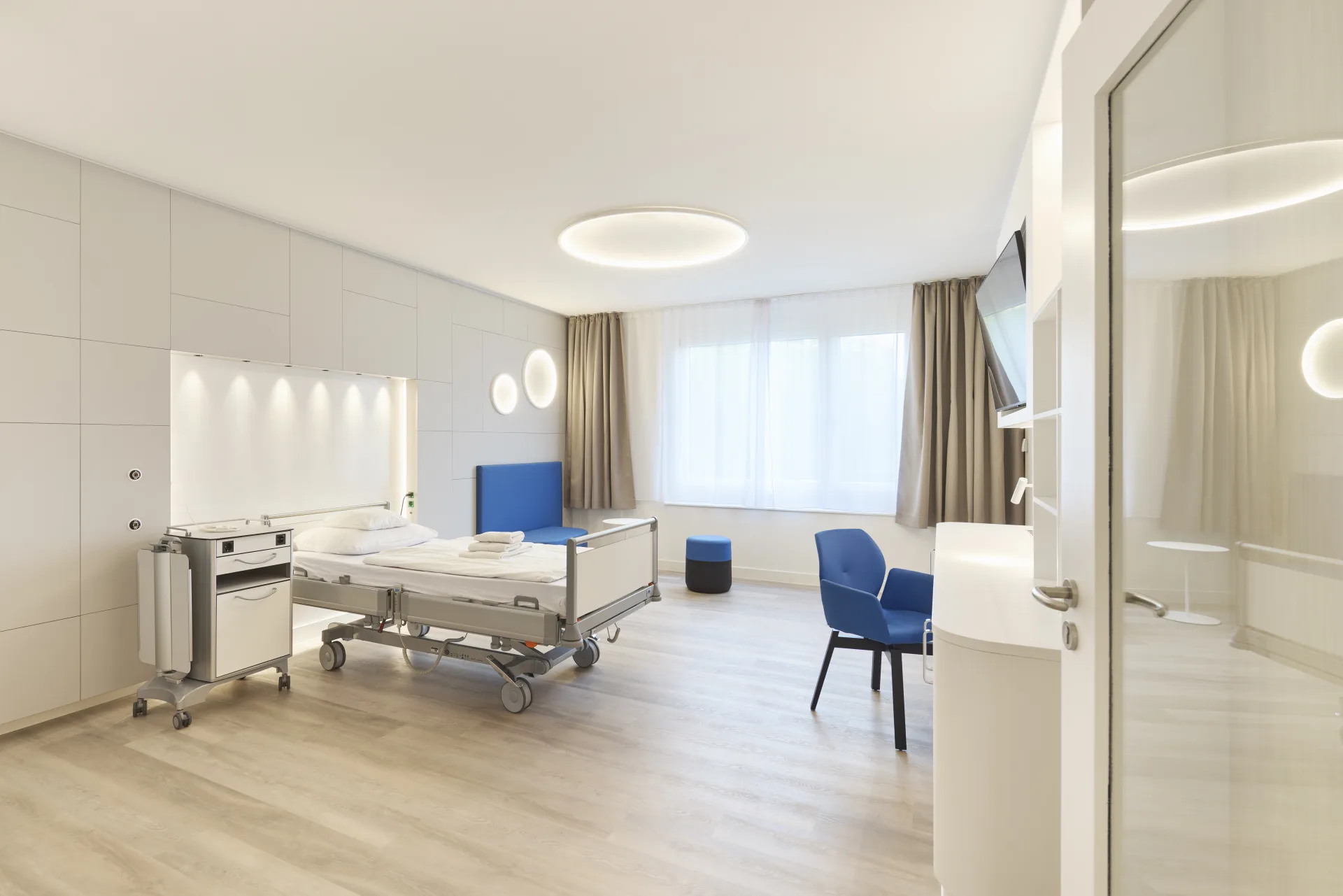 Großzügiges modernes Patientenzimmer mit Pflegebett, runder Deckenleuchte, zwei runden Wandleuchten, blauen Sesseln und hellem Holzboden