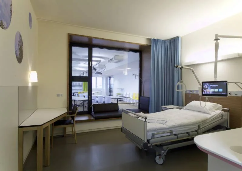 Patientenzimmer mit Pflegebett, schwenkbarem Monitor am Bett, Wand mit kreisförmigem Motiv und Fenster mit Blick in einen hellen Aufenthaltsbereich